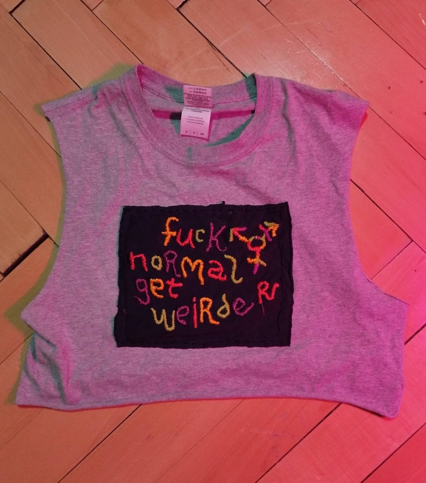 Fotka šedého trička rozstříhaného do tílkového croptopu s černým čtvercem látky s veselou nášivkou, kde stojí fuck normal, het weirder, což je v překladu mrdat normálnost, buďme divnější. Vedle textu je trans symbol. Bavlnky jsou v barvách oranžové, červené, zelené a fialové.