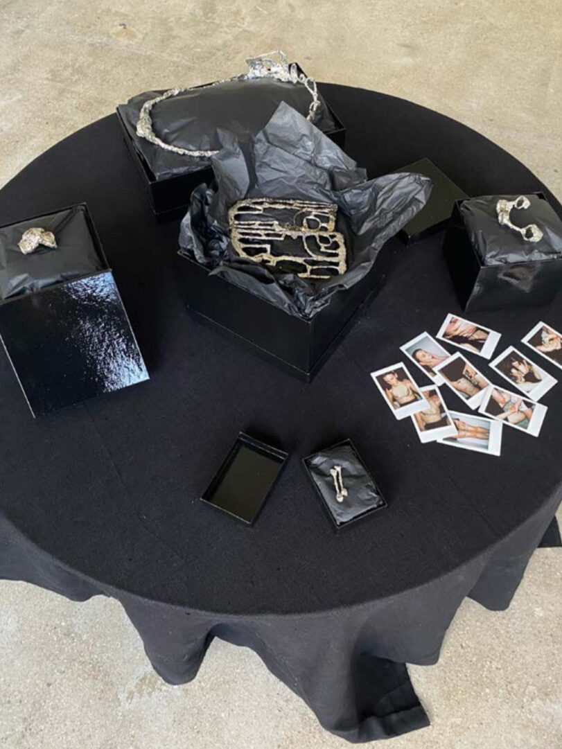 Fotka stolu s černým ubrusem a černými dárkovými krabicemi s cínovými šperky. Vedle leží polaroidové fotky.